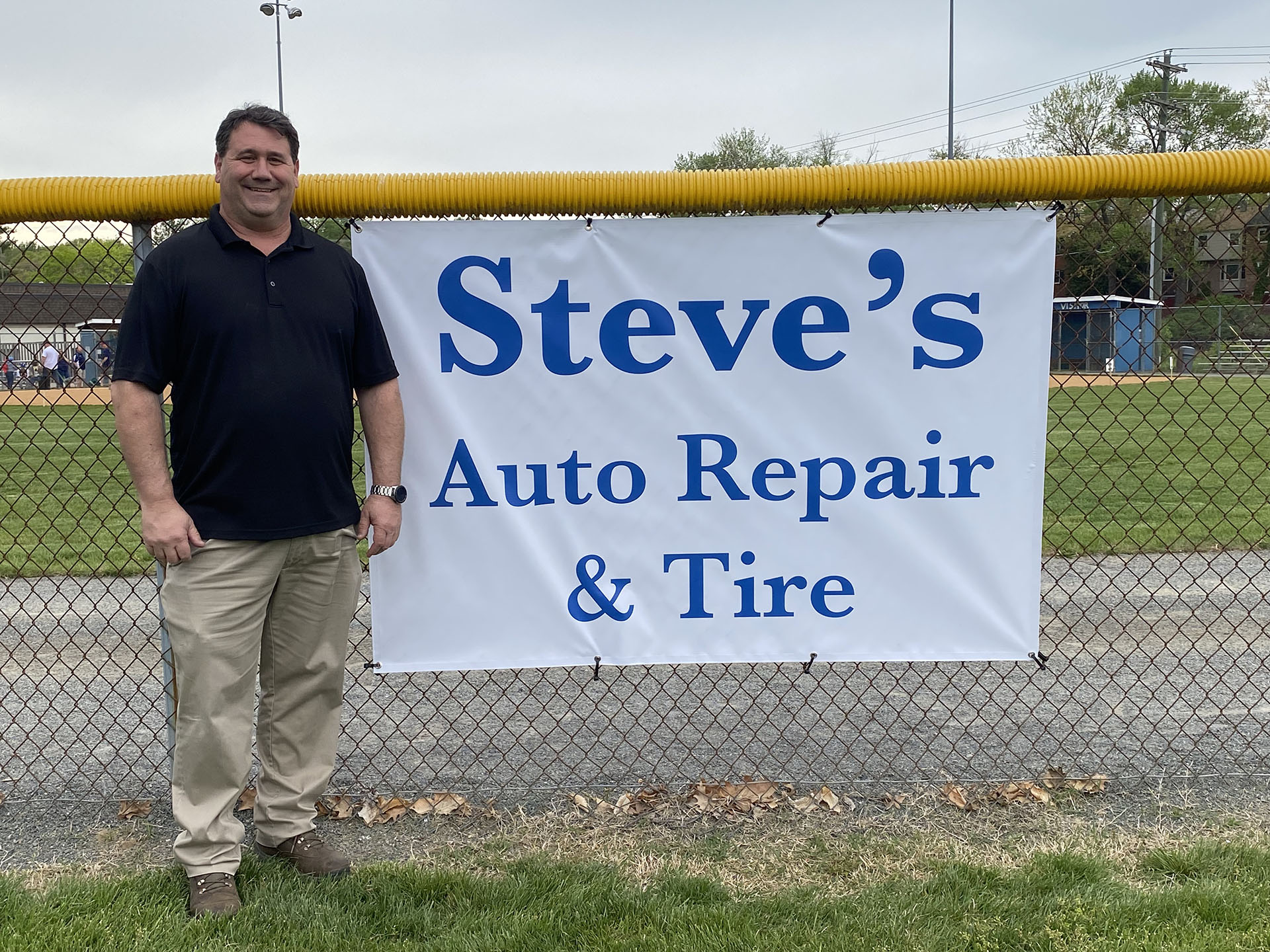 Steve's Auto Repair and Tire owner standing next to Steve's Auto Repair sign at baseball field.